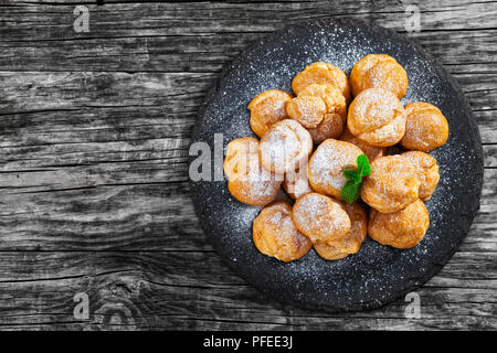 Deliziosi bignè farciti di crema sulla piastra di ardesia, spolverata di zucchero a velo, sul vecchio legno scuro tabella, vista da sopra Foto Stock