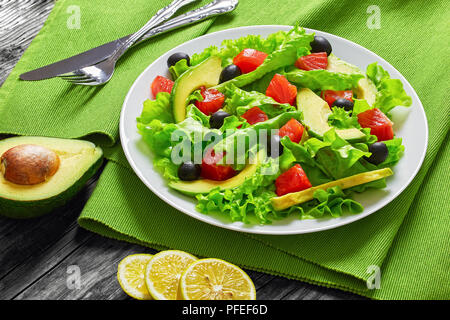 Salmone, Verde foglie di lattuga, fette di avocado e olive insalata sul piatto bianco sulla tabella mat sul vecchio dark di tavole di legno con la metà di avocado e lem Foto Stock