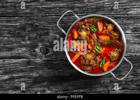 Rabo de toro o coda di vitellone stufato di carne con verdure, timo, alloro, aglio e peperoncino jalapeno pezzi nella pentola di cottura sul vecchio legno scuro tabella, tradizionale Foto Stock