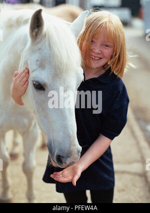 Giovane ragazza in blu in alto tenendo la testa di un cavallo bianco, altri mano sotto la bocca di cavalli. Foto Stock