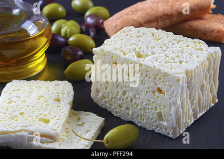 Pezzo di delizioso formaggio Feta in grigio scuro ardesia con sfondo verde e olive kalamata, сrispy salati bastoncini di pane e una bottiglia di olio di oliva, vista Foto Stock