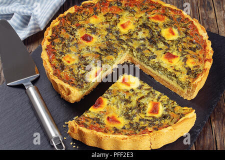 Spinaci, formaggio saporito Quiche tagliare fetta su nero ardesia, spatola e asciugamani da cucina in legno scuro, sfondo semplice ricetta, vista da sopra, close-up Foto Stock