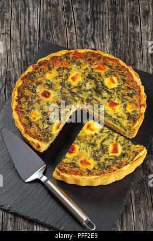 Torta salata di ricca crema di uovo, spinaci e formaggio con tagliare una fetta sul vassoio di pietra con una spatola, vista verticale dal di sopra, stile rustico Foto Stock