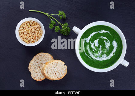 Deliziosa crema-luminoso verde zuppa di spinaci con panna acida in doppia gestite Soup Bowl sulla pietra nera piastra con i pinoli in una piccola ciotola e insieme siamo Foto Stock