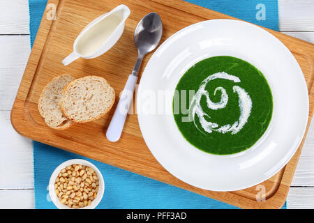 Estate a freddo una sana zuppa di spinaci, il cipollotto, broccoli in ampio cerchio piatto sul tavolo bianco, con crema e pinoli sul tagliere, vista dal Foto Stock