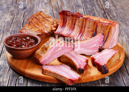 Deliziosi piatti di carne di maiale affumicata costole intere e tagliare a fette con salsa barbecue sul round tagliere in legno scuro, tabella, vista da sopra, close-up Foto Stock