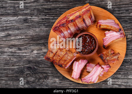 Deliziosi lastra affumicato di costolette di maiale e fette di nervatura con salsa barbecue sul tondo in legno bordo scuro sul tavolo rustico, vista da sopra Foto Stock