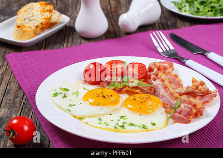 Croccante di pancetta fritta, Sunny Side fino le uova, la rucola e i pomodori sulla piastra bianca sul tavolo scuro, baguette francese toast, il sale e il pepe su backgrou Foto Stock