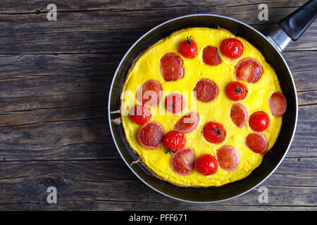 Bella Frittata con salsiccia tagliata a fettine e i pomodori ciliegia sulla padella sul tavolo di legno, vista da sopra Foto Stock