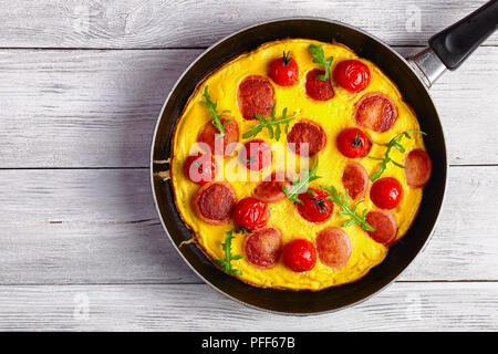 Facile e veloce prima colazione - bella frittata con salsiccia tagliata a fettine, interi di pomodori ciliegini e rucola fresca su una padella sul tavolo di legno, vista fr Foto Stock