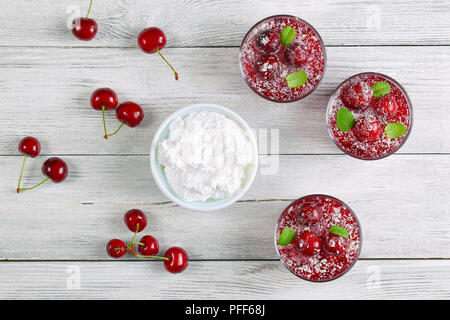 Deliziosi dessert estivo - Gelatina di ciliegia in tre tazze di vetro decorate con polvere di noce di cocco e le foglie di menta sul tavolo di legno con panna montata nel recipiente, Foto Stock