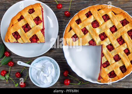 Fetta di in casa di ciliege acide la teglia sulla piastra e tutta la torta tagliata a fette in una teglia su dark tavolo in legno con la panna montata in un recipiente e freschi maturi Foto Stock