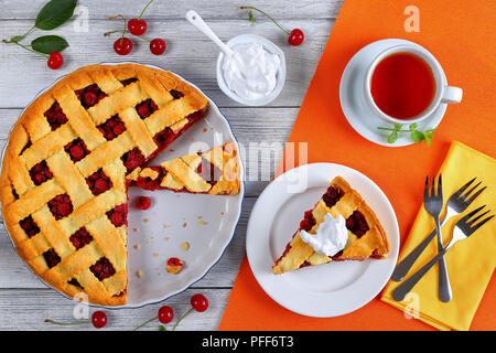 Fetta di deliziosa casa di ciliege acide pie sulla piastra. Ciotola con panna, dessert forcelle e tart tagliato a fettine e la tazza di tè sul tavolo di legno, cl Foto Stock