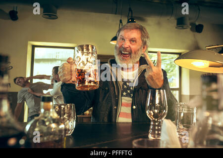 Senior uomo barbuto di bere alcolici in pub e la visione di un programma di sport in TV. Godendo di birra. Uomo con la tazza di birra seduti al tavolo. Calcio o dello sport ventola. Lotta di ventilatori in background Foto Stock