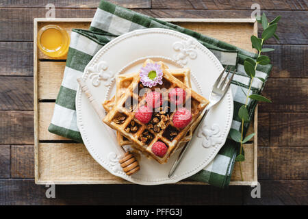 Cialde belghe con miele e lamponi per la prima colazione. Cialde decorate con fiori, frutti di bosco, muesli e del miele per servire in legno vassoio. Vista superiore Foto Stock