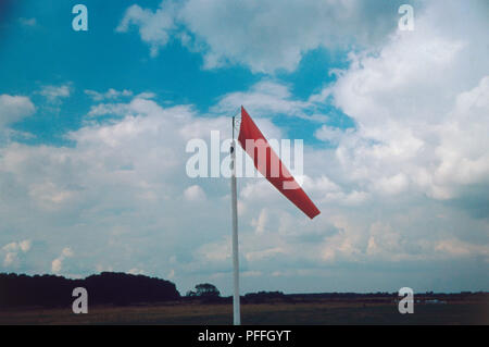 Antivento rosso su un campo di aviazione, essendo leggermente sollevato leggermente dal vento leggero, il bianco delle nuvole in cielo blu, campo e alberi in background. Foto Stock