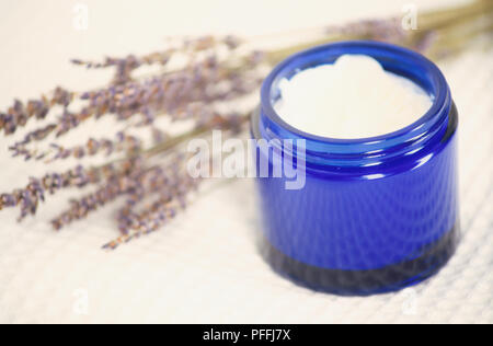 Blue il vasetto di vetro contenente crema lavanda, offuscata, essiccati fiori di lavanda in background, vicino, vista in elevazione. Foto Stock