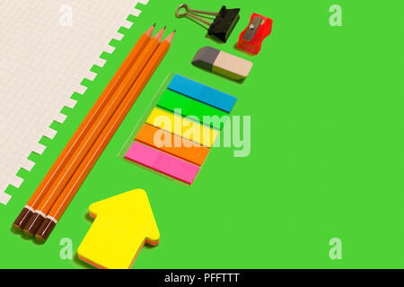 Foglio di carta bianco, matite, adesivi, affilatrice, eraser e una sovrapposizione in diagonale giacente su di una superficie verde. concetto di cancelleria educativa. spa gratis Foto Stock