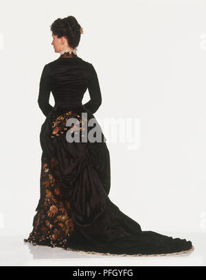 Una donna che indossa un tardo XIX Secolo trambusto di seta abiti di colore scuro con disegni floreali, vista da dietro Foto Stock