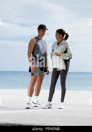 L uomo nel serbatoio superiore, pantaloncini, cappello da baseball, rivolta verso la donna in leggings e camicia bianca tenendo la bottiglia di acqua Foto Stock