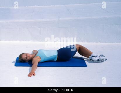 La donna nel serbatoio superiore e pantaloncini giacente sul retro sul tappeto blu, bracci giacenti dritto fuori in corrispondenza dei due lati, hips ritorto per un lato, esecuzione di rullo laterale Foto Stock