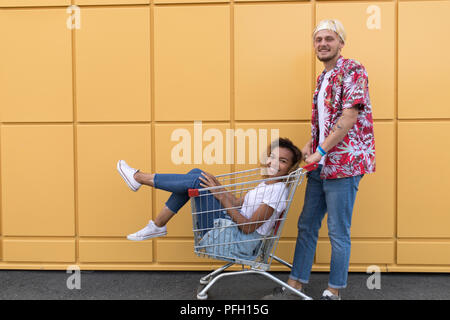 Immagine di giovani amici sorridente su parete gialla. Buon divertimento con il carrello della spesa. Foto Stock