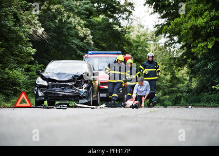 I vigili del fuoco in esecuzione per il soccorso di una donna giaceva incosciente sulla strada dopo un incidente di automobile. Un uomo maturo tenendo la sua mano controllando il suo impulso. Copia spa Foto Stock