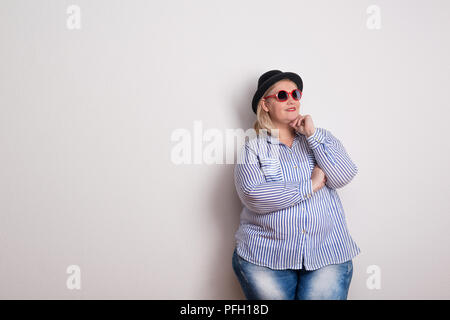 Ritratto di un attraente Donna sovrappeso con un cappello nero e rosso occhiali da sole in un studio. Close up. Foto Stock