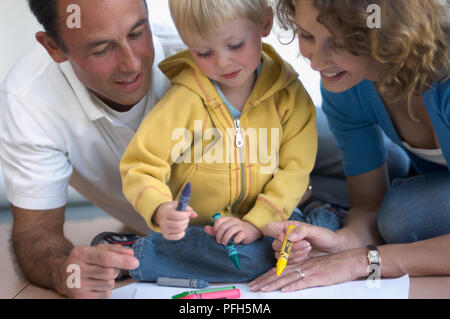 Ragazzo tenendo il blu e il verde pastelli su pezzo di carta, l uomo e la donna accovacciata accanto a lui, donna azienda crayon giallo Foto Stock