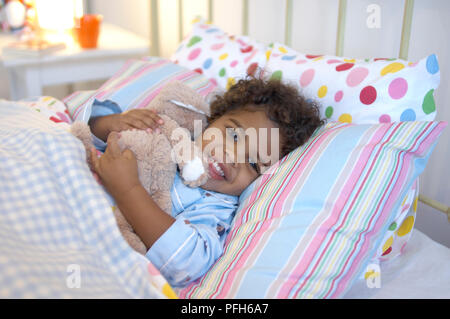 Ragazzo disteso sul letto abbracciando giocattolo imbottito, sorridente, ritratto Foto Stock