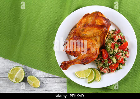 La metà di deliziose grigliate di succosa il pollo con la golden crosta marrone servita con fettine di lime e cuscus insalata di verdure sulla piastra bianca sul tavolo verde ma Foto Stock