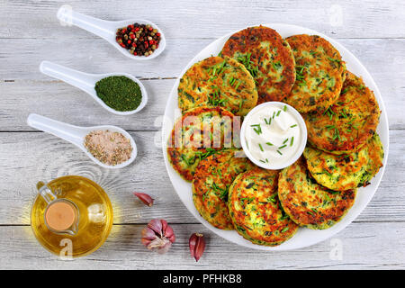 Deliziose frittelle di zucchine su piastra bianca con panna acida nel centro del piatto spolverati con un trito di erba cipollina, spezie. Bottiglia di Olio su legno Foto Stock