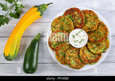 Deliziose frittelle di zucchine su piastra bianca con panna acida nel recipiente nel centro del piatto spolverati con un trito di erba cipollina, materie le zucchine sul t di legno Foto Stock