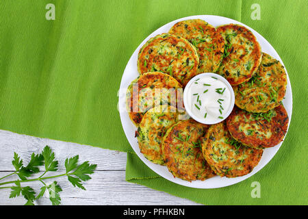 Deliziose frittelle di zucchine su piastra bianca con panna acida nel recipiente nel centro del piatto spolverati con un trito di erba cipollina, vista dall'alto Foto Stock