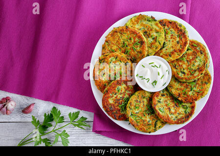 Deliziose frittelle di zucchine su piastra bianca con panna acida nel recipiente nel centro del piatto spolverati con un trito di erba cipollina, vegetariano sano destinatario Foto Stock