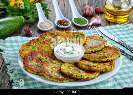 Deliziose frittelle di zucchine cosparso con un trito di erba cipollina sul piatto di portata con panna acida, sul vecchio dark tavolo in legno con ingredienti su sfondo, Foto Stock