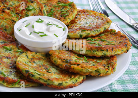 Close-up di deliziose frittelle di zucchine cosparso con un trito di erba cipollina sul piatto di portata con panna acida, sul vecchio dark tavolo in legno con ingredienti su Foto Stock