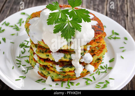 Croccanti frittelle di zucchine cosparso con un trito di erba cipollina sul piatto di portata con panna acida, sul vecchio legno scuro tabella, vegetariane ricetta, vista orizzontale Foto Stock