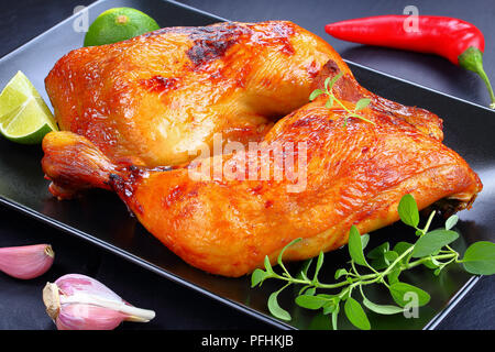 Delizioso pollo arrosto con gambe dorate e croccanti in pelle marrone con le fette di limone e fresco verde di foglie di timo sulla piastra nera sul vassoio di ardesia, vista da ab Foto Stock