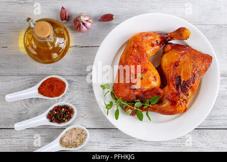 Deliziosi arrosti di cosce di pollo con croccante dorato marrone pelle con fresco verde di foglie di timo sulla piastra bianca, sul tavolo di legno con spezie e bot Foto Stock