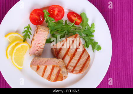 Deliziose grigliate di bistecca di tonno con strisce servita con foglie di rucola insalata di pomodori e le fette di limone sulla piastra bianca sulla tabella mat, di facile e rapida destinatario Foto Stock