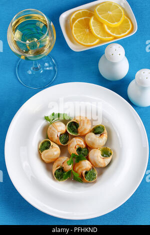 Porzione di Escargots de Bourgogne o lumache con erbe aromatiche, burro, aglio sulla piastra bianca sulla tabella MAT con un bicchiere di vino bianco, le fette di limone, verticale vi Foto Stock