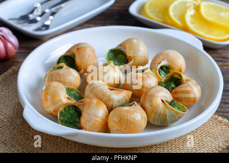 Cucina francese - Escargots de Bourgogne o lumache con erbe aromatiche, burro, aglio sulla piastra bianca con forche, le fette di limone.,l'aglio sul tavolo, vista orizzontale Foto Stock