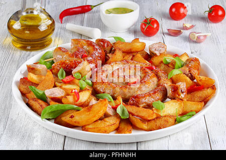 Salsicce bavaresi - fritte salsicce a spirale sul piatto bianco con spicchi di patate pezzi di peperoncino e le foglie di basilico, sul tavolo di legno con aglio e olio d'oliva oi Foto Stock