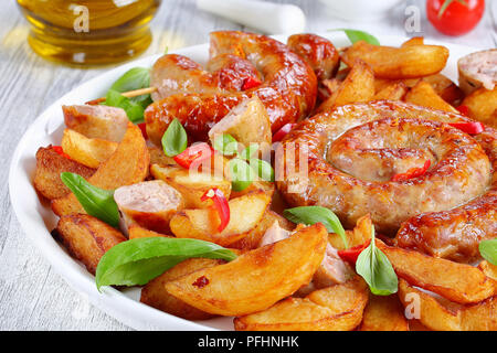 Salsicce bavaresi - fritte salsicce a spirale sul piatto bianco con spicchi di patate pezzi di peperoncino e le foglie di basilico, sul tavolo di legno con aglio e olio d'oliva oi Foto Stock