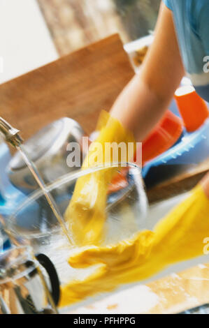 Donna di colore giallo che indossa guanti di gomma lavaggio ciotola di vetro sotto acqua corrente nel lavello da cucina, vista laterale. Foto Stock