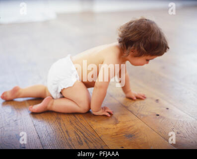 Ragazza toddler in bianco pannolino strisciare lungo il pavimento in legno e vista laterale Foto Stock