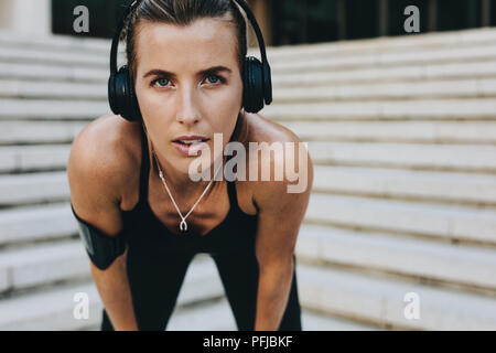 donna in forma fisica che si allena all'aperto piegandosi in avanti con le mani sulle ginocchia. donna in forma fisica che indossa le cuffie wireless per rilassarsi durante l'allenamento. Foto Stock