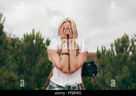 Viaggiatore femmina che copre il volto con le mani in piedi in una foresta. Donna su una vacanza portando una borsa da viaggio. Foto Stock