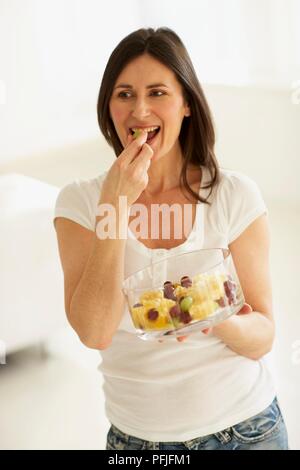 La donna in piedi e mangiare la frutta dal vaso di vetro tenuto in mano Foto Stock